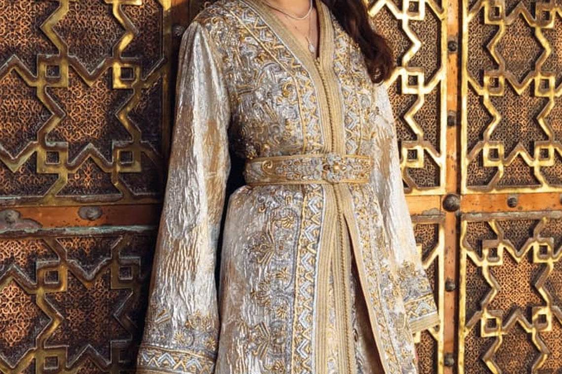 Pearl Gate ivory and silver embroidered Moroccan kaftan with a beaded belt, before an engraved brass door